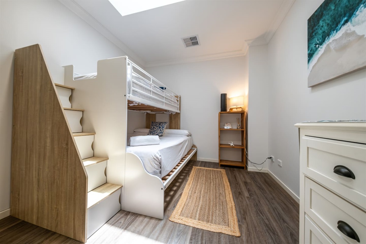 Bright and cosy third bedroom featuring a stylish bunk bed with built-in storage stairs, wood flooring, a natural woven rug, and a softly lit bookshelf in the corner.