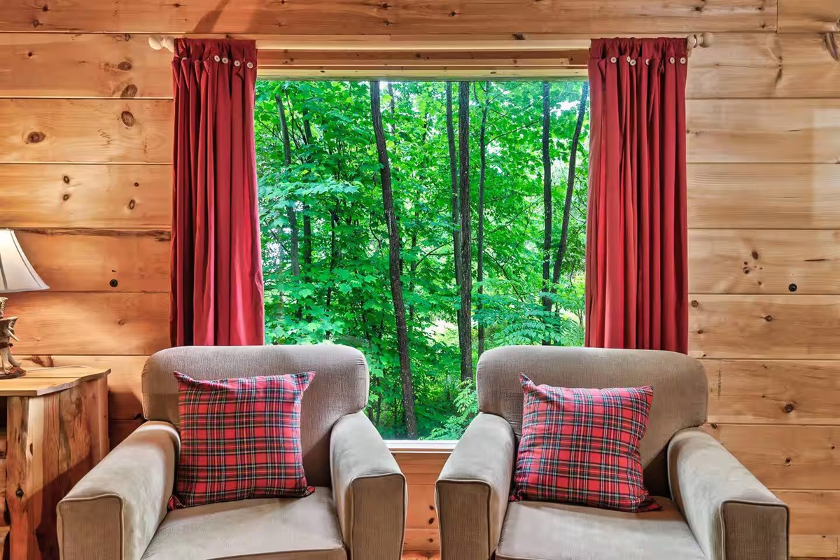 A serene seating area with plush chairs and cheerful plaid accents is complemented by a window framing the vibrant greenery, perfect for sipping coffee and enjoying the natural surroundings.