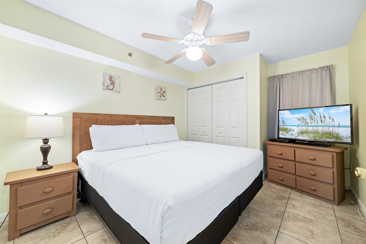 This well-appointed primary bedroom features a king-size bed with a decorative headboard, crisp white bedding, ceiling fan and nightstand with lamp, practical storage solution, and tile flooring for easy maintenance.