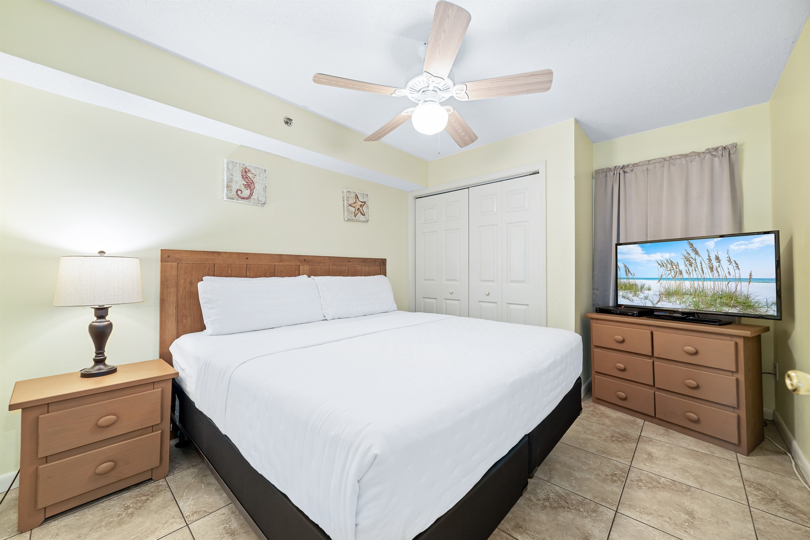 This well-appointed primary bedroom features a king-size bed with a decorative headboard, crisp white bedding, ceiling fan and nightstand with lamp, practical storage solution, and tile flooring for easy maintenance.