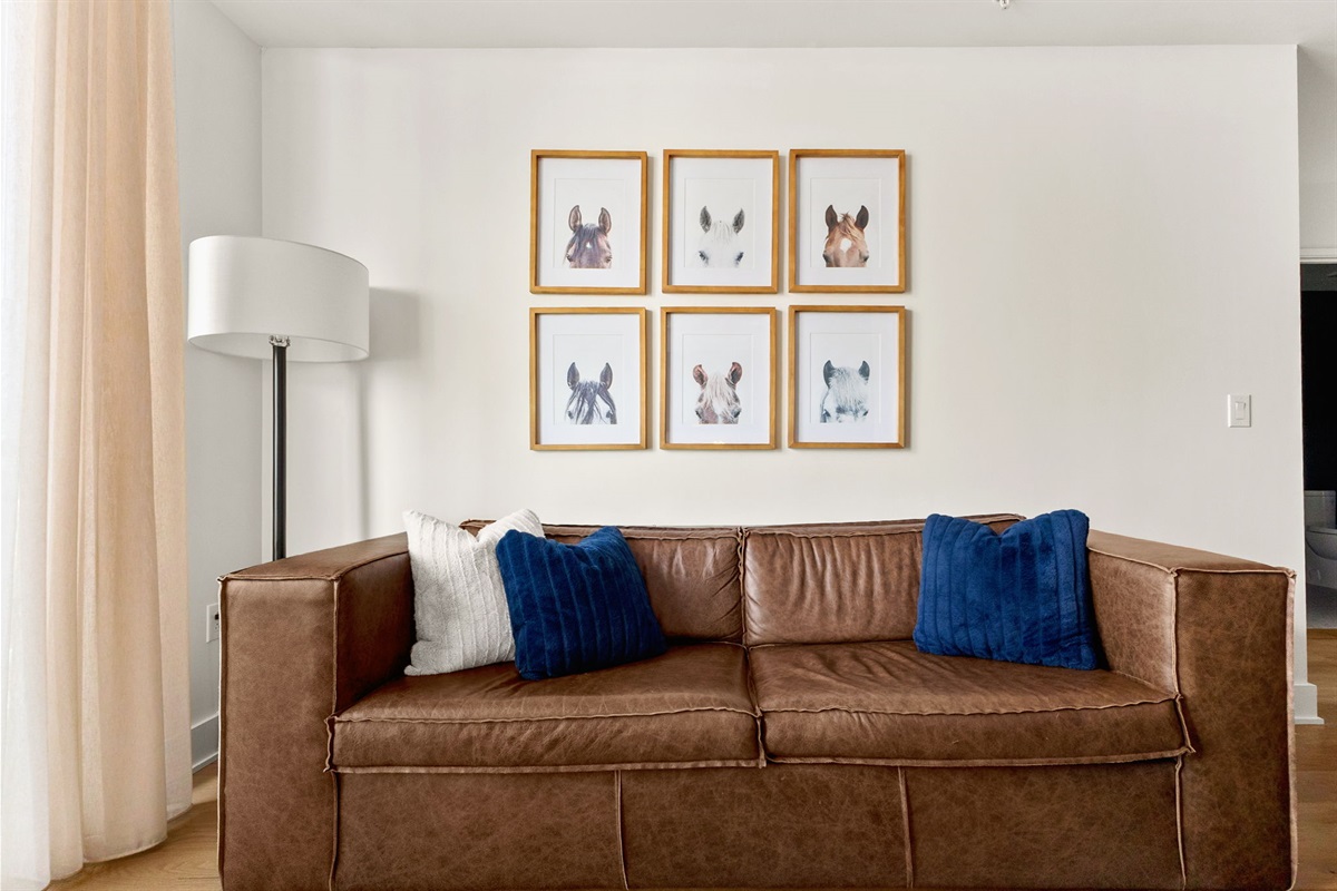 A cozy living area featuring a comfortable brown leather sofa adorned with dark blue and white pillows, centered beneath a grid of six framed horse portraits, creating a warm and stylish focal point.