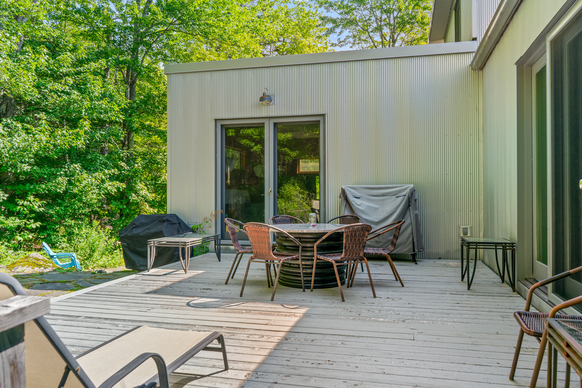 The main outdoor living space at "The Crag." This spacious deck features a circular dining setup, sun lounger, covered grill, and direct access to both the primary suite and main lounge—all bordering Acadia National Park.