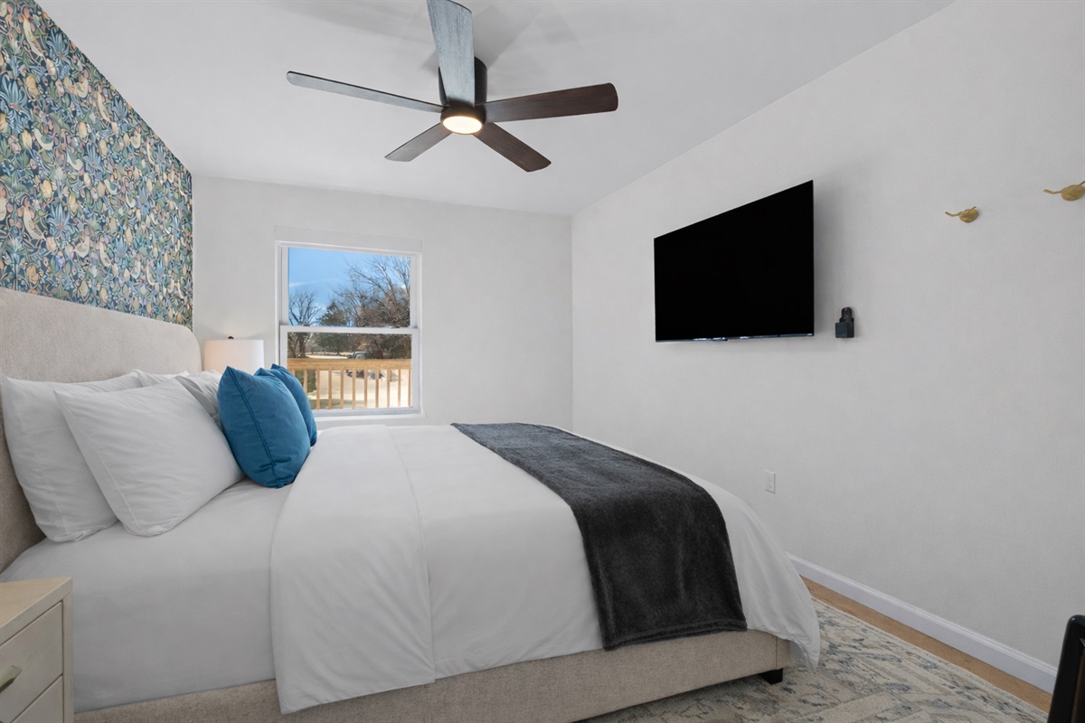 Relax in this bright bedroom featuring a plush bed, smart TV, ceiling fan, and peaceful natural light—perfect for restful nights and easy mornings.