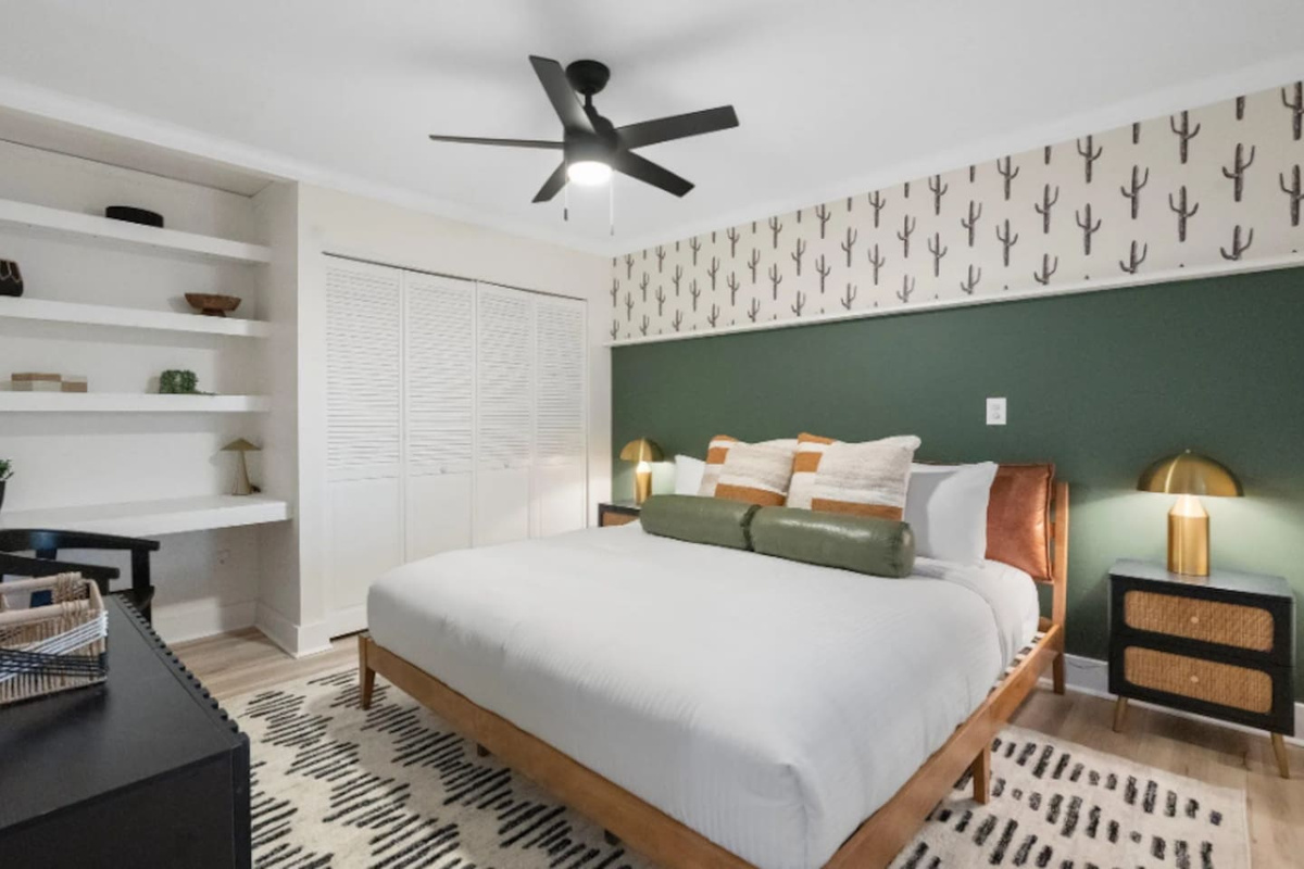 A deep green accent wall and hand-illustrated cactus print set a moody, grounded tone, while the built-in shelving and desk nook add a quiet workspace for anyone who needs one. Brass lamps warm up the whole corner.
