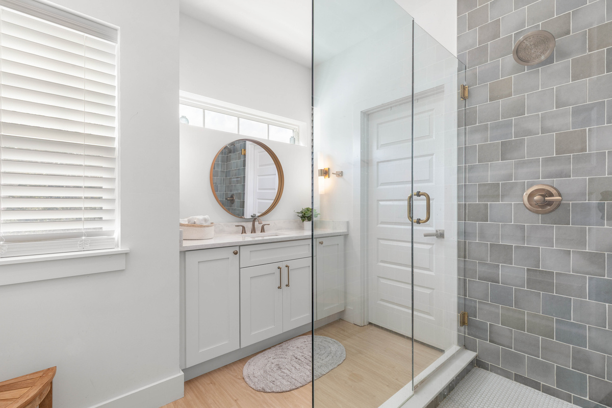 Sea-glass tile in blues and greys lines the shower, and the glass door keeps the light moving through. The vanity sits opposite with a round mirror, so two people can get ready without having to take turns.