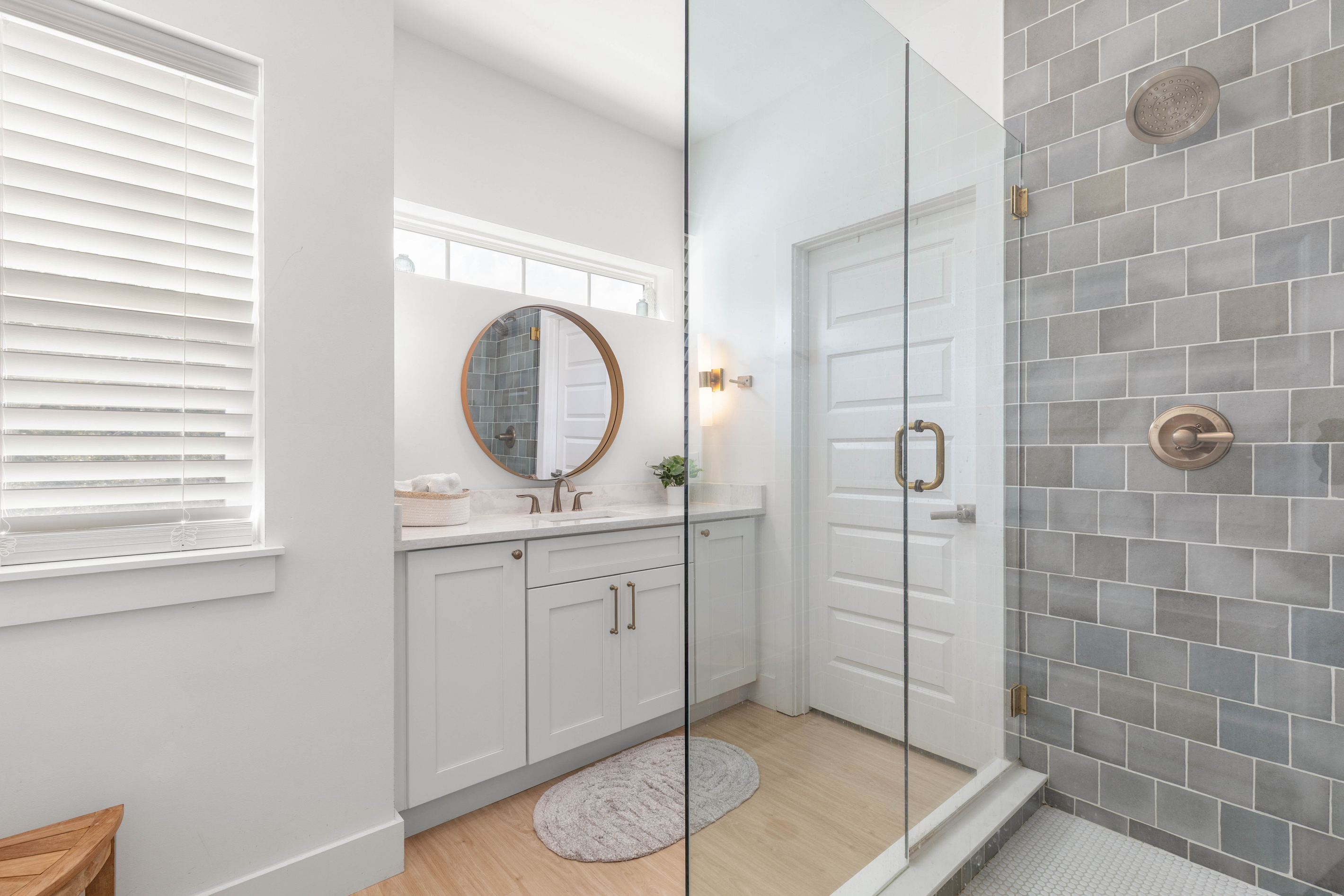 Sea-glass tile in blues and greys lines the shower, and the glass door keeps the light moving through. The vanity sits opposite with a round mirror, so two people can get ready without having to take turns.