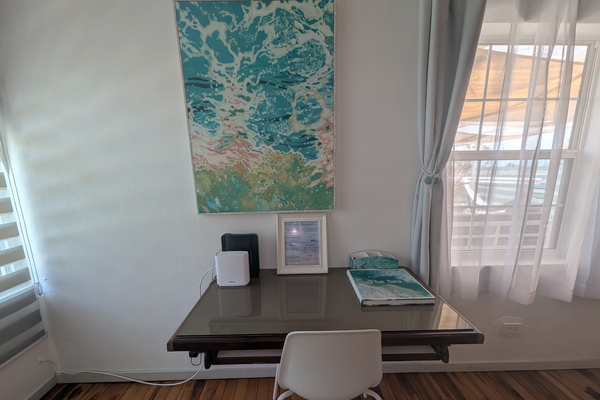 Dedicated remote workspace at Villa Coral featuring a glass-top desk, white modern chair, high-speed Wi-Fi router mesh pod, coastal wave art, and natural window lighting in Dorado, Puerto Rico.
