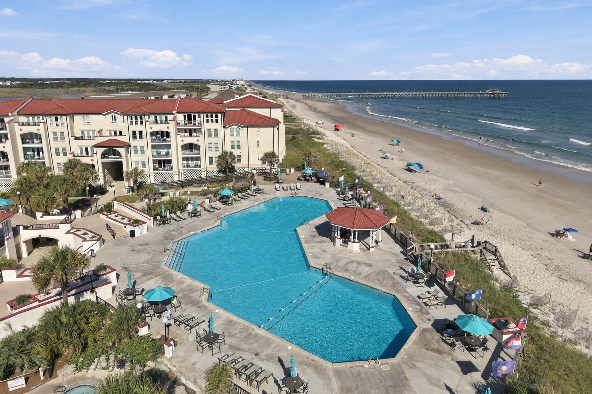 Elevated view of the outdoor pool area, featuring a swimming basin, shaded gazebo, surrounding sun deck, dune-line boardwalk, and nearby pier extending into the ocean.   