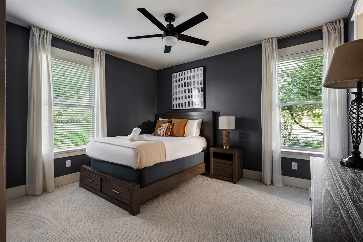 Bright and cozy bedroom with a queen bed, ceiling fan, and two sunlit windows.