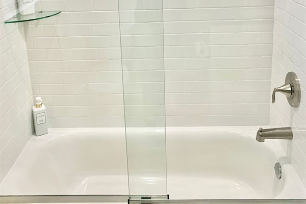 Enjoy a relaxing soak in the clean, white bathtub featuring modern brushed nickel fixtures. The space is beautifully finished with classic white subway tile and a sleek glass door, providing a bright and polished feel.