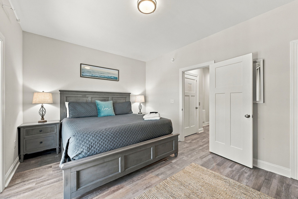 Primary master bedroom view showcasing the dark grey wooden king bed, coastal artwork, wood-look flooring, and an open doorway leading to the ensuite bathroom.   