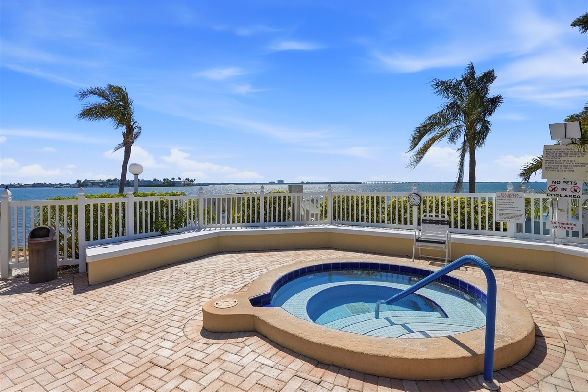 The resort-style heated pool is surrounded by palm trees, lounge chairs, and beautifully maintained grounds, all set against the scenic bay area backdrop.