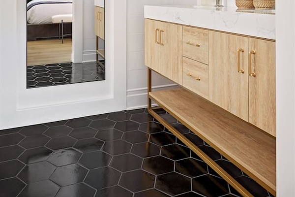 A spacious second-floor ensuite bathroom features modern hexagon tile floors and a large white oak dual-sink vanity with high-end polished nickel Kohler faucets. Custom designer lighting and a full-length mirror complete an elegant, spa-like retreat.