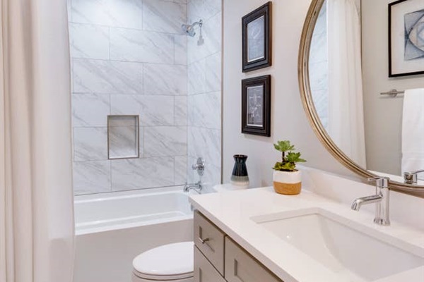 - White vanity with quartz top and brushed nickel fixtures offers a spa like feel - Full tub with marble style tile and inset shelf perfect for a relaxing soak - Round mirror, framed art, and fresh greenery complete the calming design  