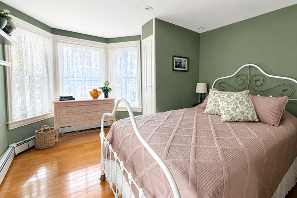 A queen bedroom on the second floor with original hardwood floors, a bay window dressed in lace, and that particular quiet that only old houses have.