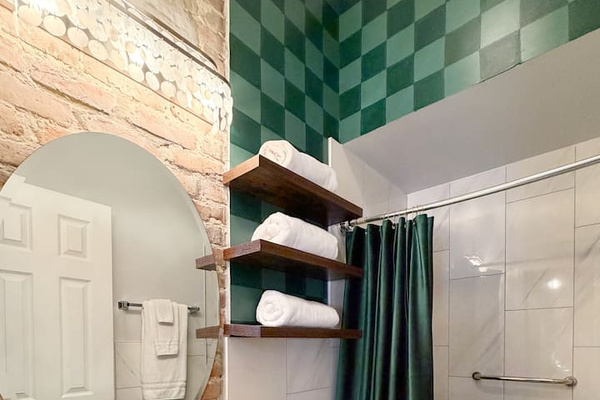 Accessible from the whole house via the kitchen, the hallway bathroom features exposed brick, emerald checkerboard walls, and spa-style walk-in  shower. Fresh towels and essentials provided for a comfortable New Orleans stay. 