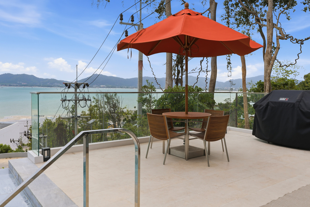 Terrace with red umbrella, dining table, ocean and mountain views beyond glass railing.