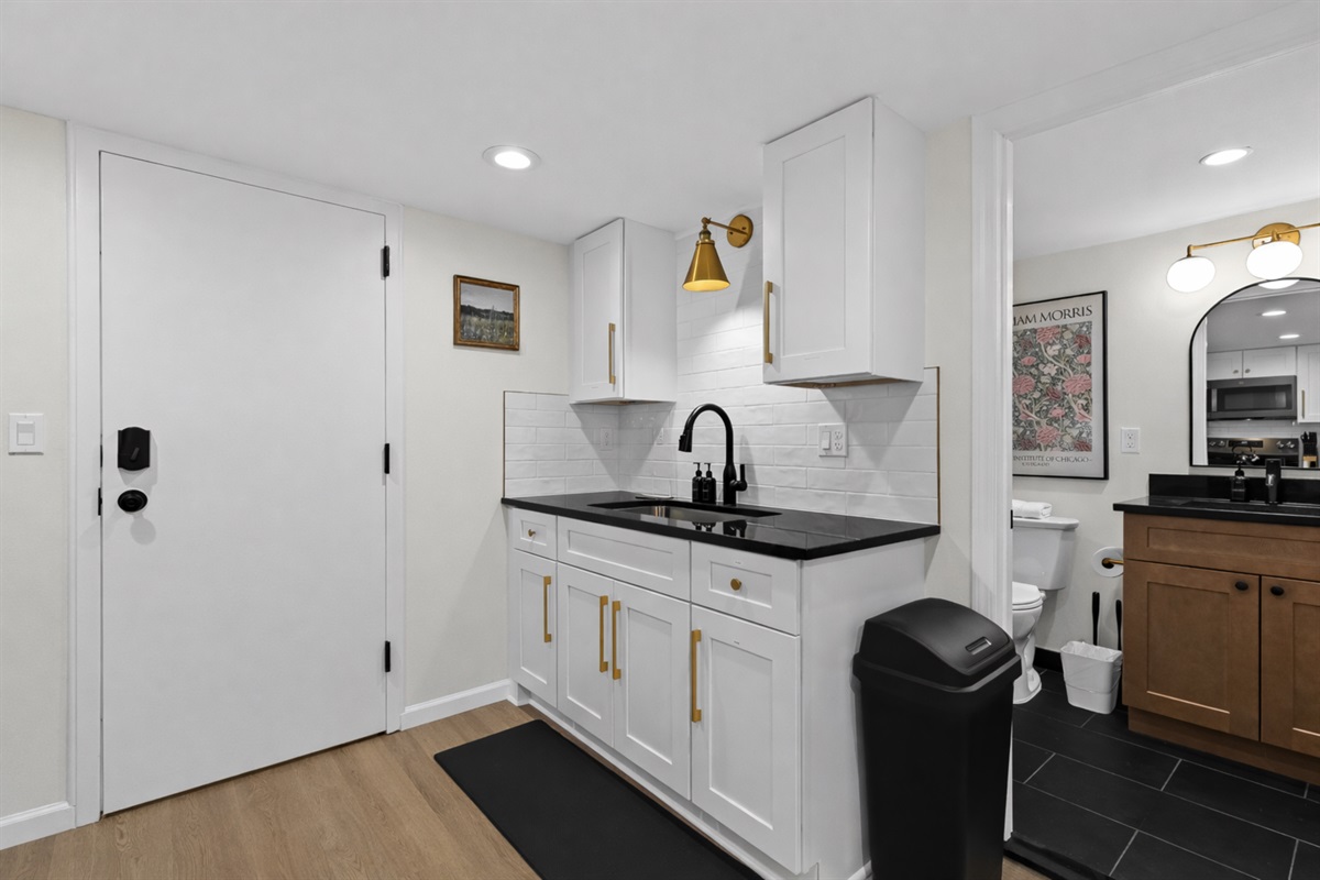 Stylish kitchenette and bath access with crisp white cabinetry, sleek black counters, and brass accents—designed for convenience, comfort, and a polished stay.