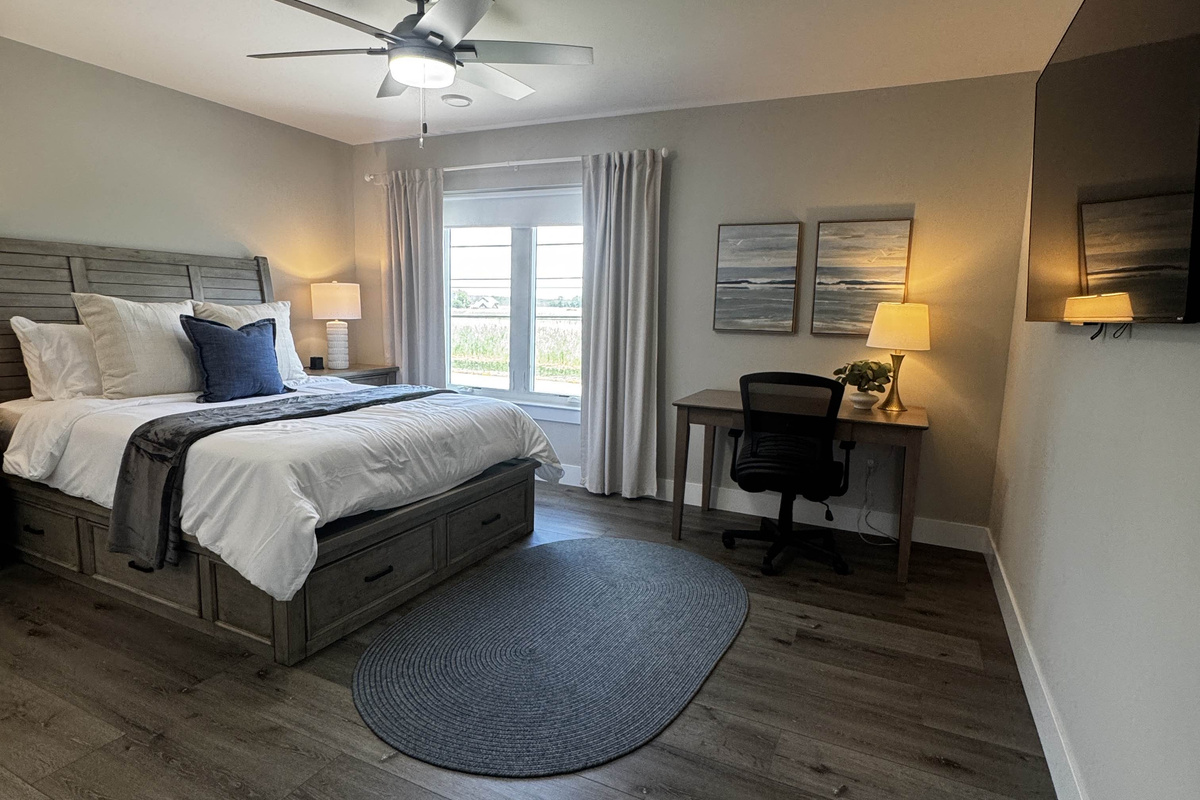 Bright and comfortable bedroom featuring a queen bed, dedicated workspace, and soft neutral décor. Large windows bring in natural light and offer peaceful outdoor views, creating a calm and inviting space for rest or remote work.
