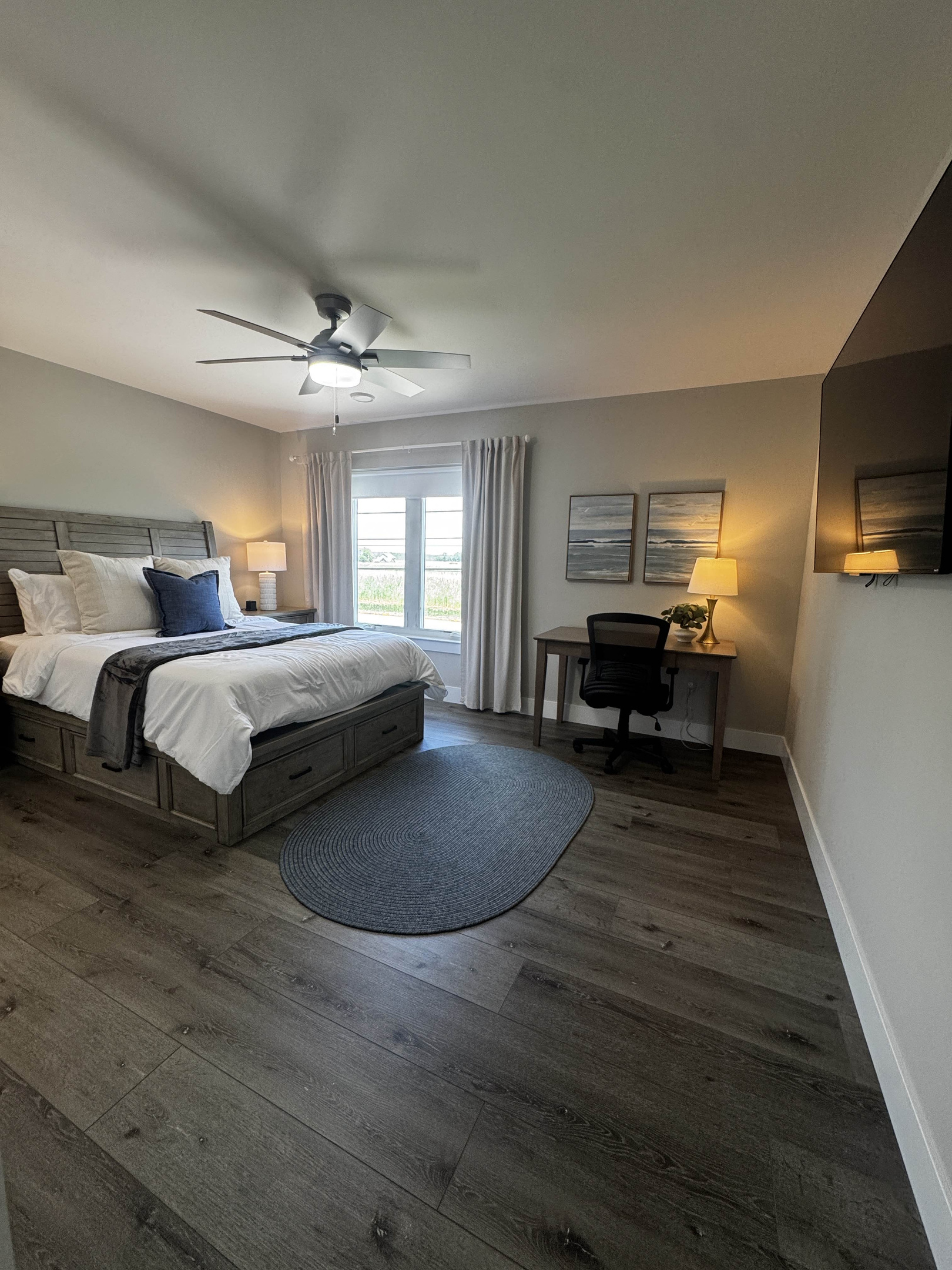 Bright and comfortable bedroom featuring a queen bed, dedicated workspace, and soft neutral décor. Large windows bring in natural light and offer peaceful outdoor views, creating a calm and inviting space for rest or remote work.