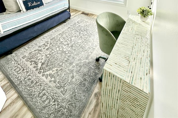 This functional nook features a textured white console desk with a sage green velvet chair. The space is anchored by a large, gray patterned rug over wood-look flooring and sits adjacent to a daybed with a pull-out trundle.