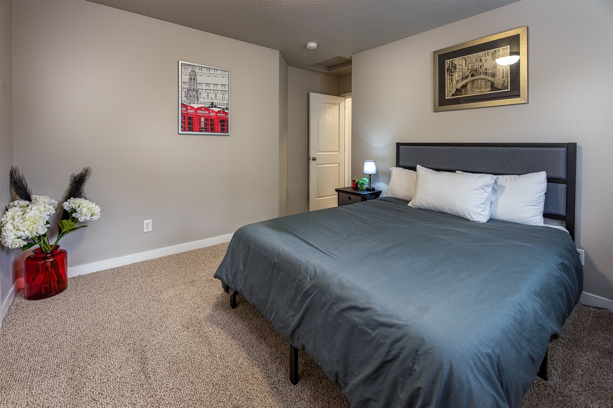 A cozy bedroom featuring a queen-sized bed with gray bedding, two white pillows, a bedside table with a lamp and small decor, a vibrant red vase with white flowers, a framed wall art depicting a London scene, and a door leading to a bathroom or closet.