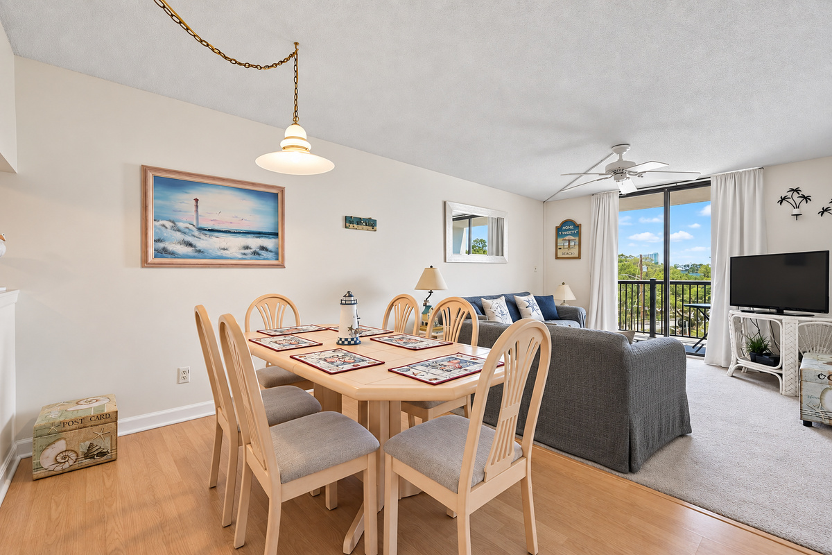 Dining area with seating for six, beach-style decor, and an open view into the living room and balcony.