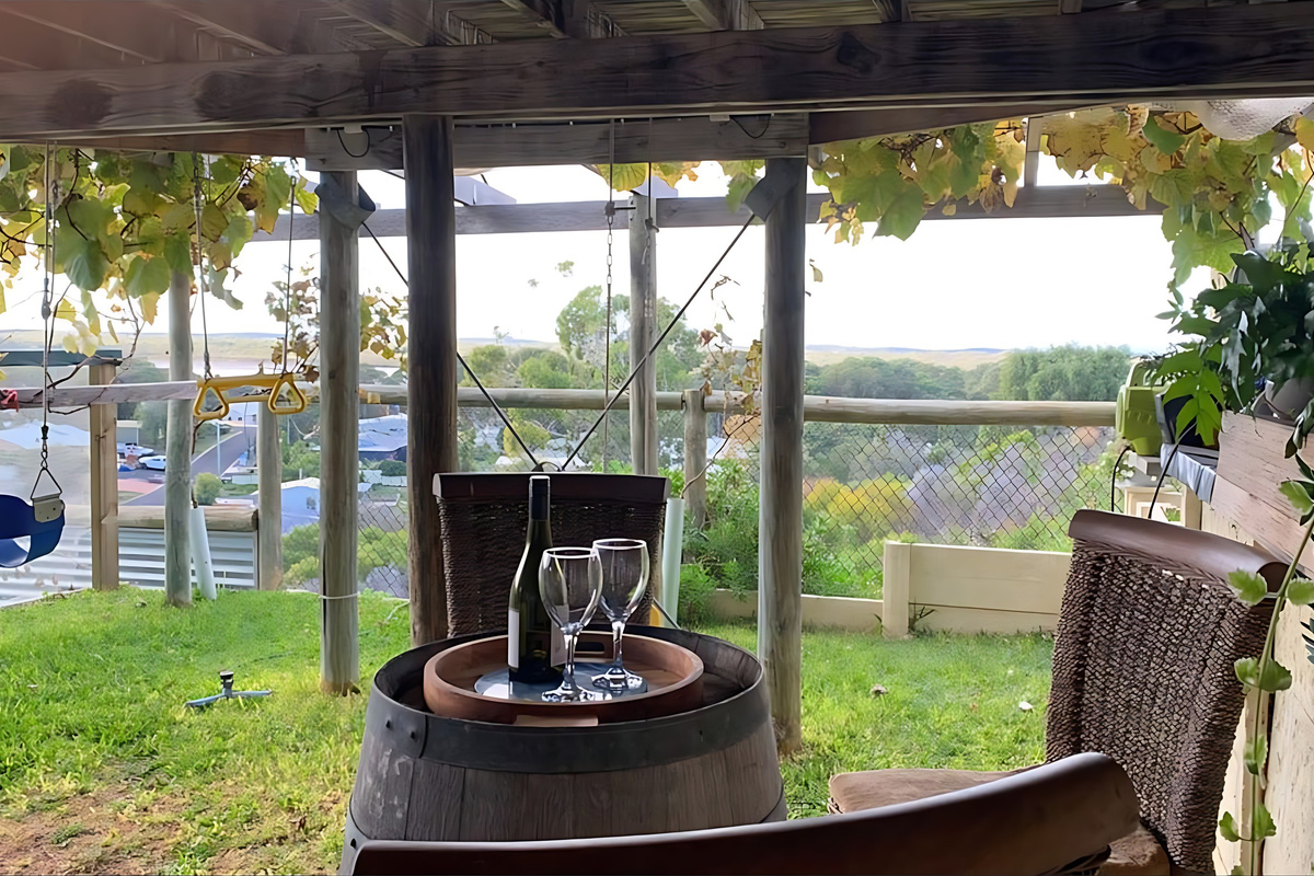 A vine-shaded seating area in the backyard has a wine-barrel table and wicker chairs, with a baby swing and gym rings nearby and a view across Preston Beach to Lake Preston.