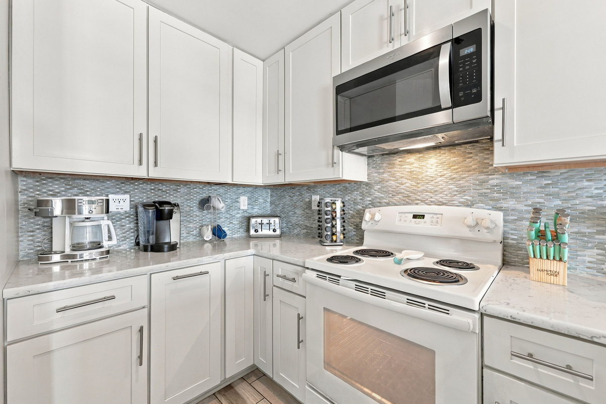 Detailed perspective of the fully equipped kitchen prep area, highlighting the white shaker-style cabinetry, shimmering glass mosaic backsplash, smooth white quartz counters, and integrated stainless steel microwave above the electric range. 