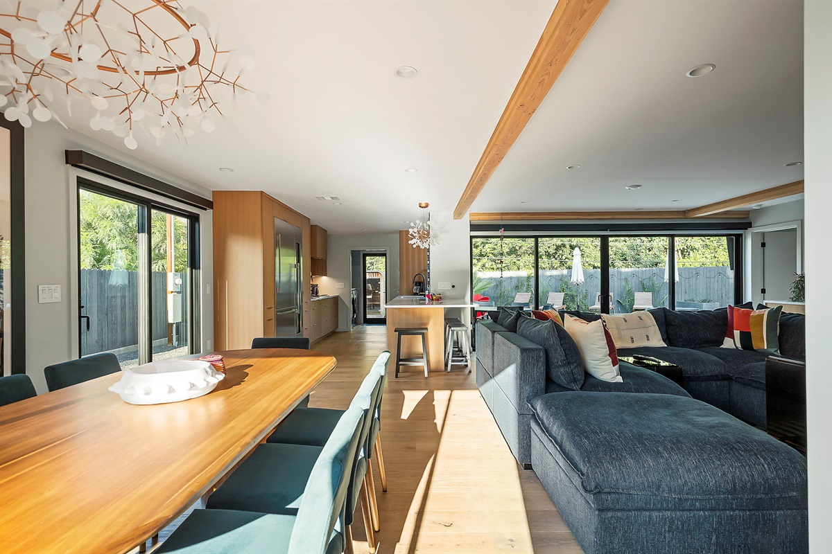 A bright and open living and dining area featuring a long wooden table, a large sectional sofa, and a modern kitchen with light wood cabinetry, all connected by seamless hardwood flooring and exposed beams.