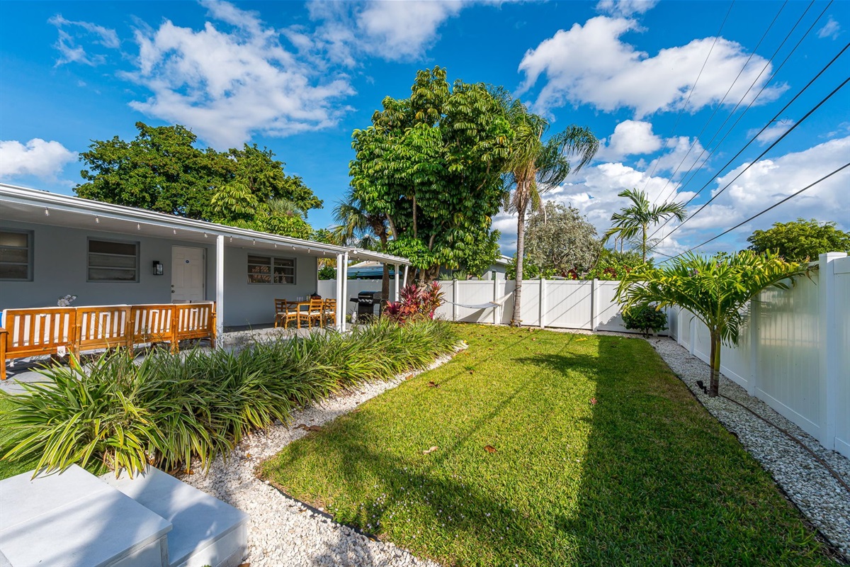 A serene covered patio with chic seating, alfresco dining, a gas grill, and a two-person hammock, all framed by a mix of manicured turf and lush lawn—your private outdoor retreat.
