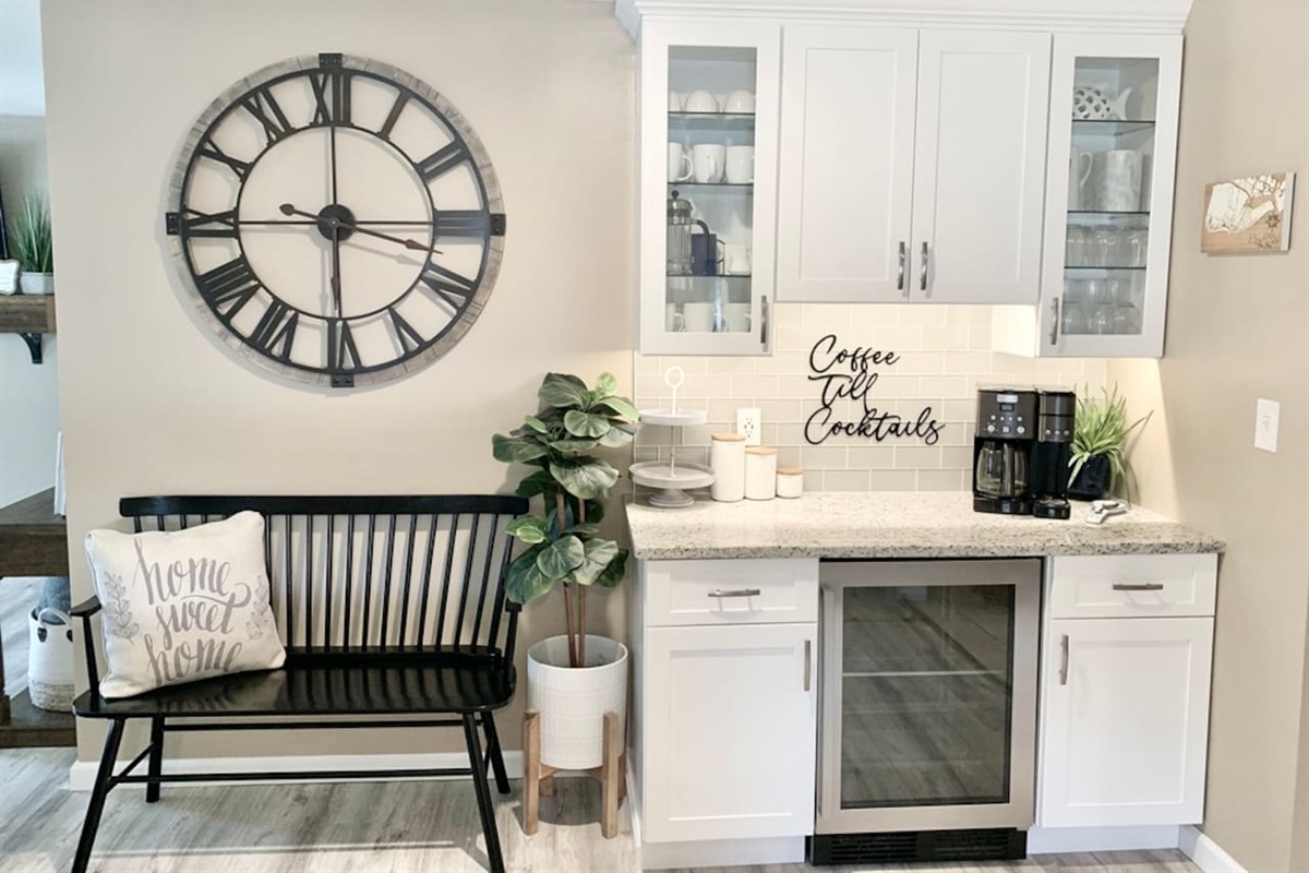 Start your morning or end your evening at our dedicated beverage bar. Features a drip coffee maker, wine cooler, and "Coffee Till Cocktails" decor. A charming black bench and oversized wall clock complete this stylish and functional nook.