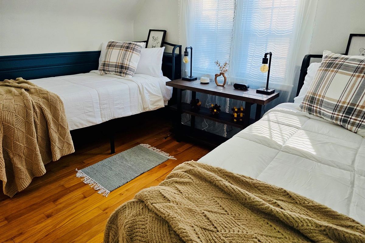 Twin XL beds with cozy lighting, charging stations, alarm clock, and white noise machine offer restful nights on rich hardwood floors. A wall-mounted TV (not pictured) adds entertainment, and an optional safety rail available for added peace of mind.