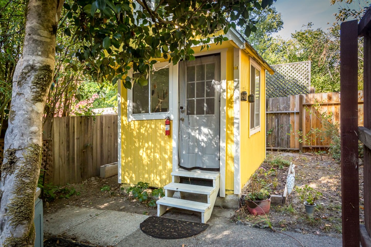 - Charming yellow garden cottage with white trim and front steps
- Surrounded by greenery and privacy fencing for a peaceful escape
- Perfect for creative projects, remote work, or quiet relaxation
