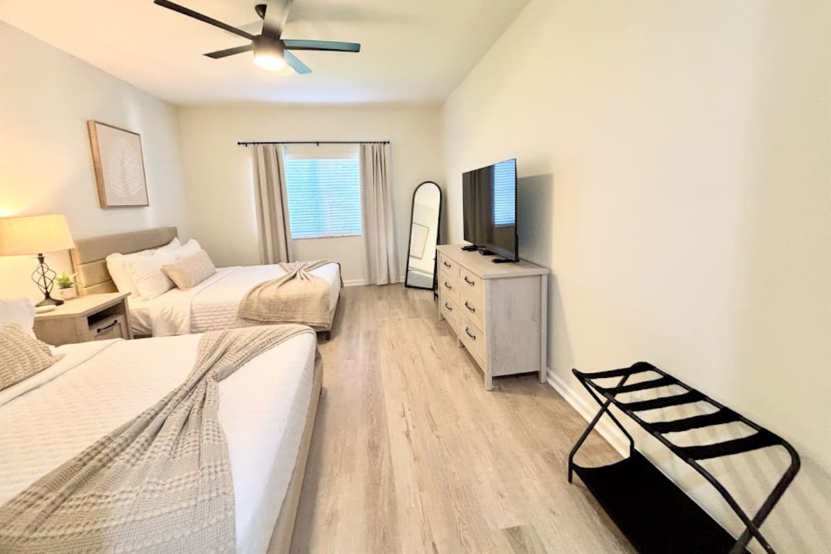 Spacious bedroom with two comfortable beds, wood-look flooring, ceiling fan, TV, dresser storage, and a convenient luggage rack.