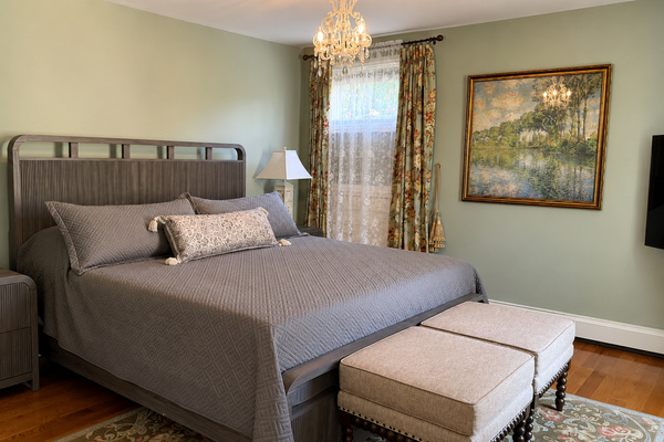 The primary suite — a king bed, crystal chandelier, hardwood floors, and an en suite bath. The floral drapes and lace sheers are a nice nod to the Victorian bones of the house.