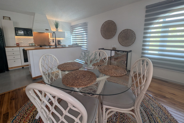 Upstairs dining area at Villa Coral featuring a round glass-top table, white rattan chairs, woven placemats, a colorful jute rug, and a clear view of the adjacent kitchen island in Dorado, Puerto Rico.
