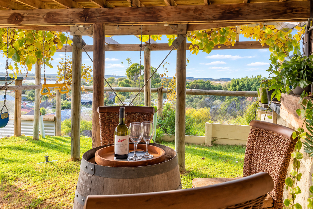 A vine-shaded seating area in the backyard has a wine-barrel table and wicker chairs, with a baby swing and gym rings nearby and a view across Preston Beach to Lake Preston.