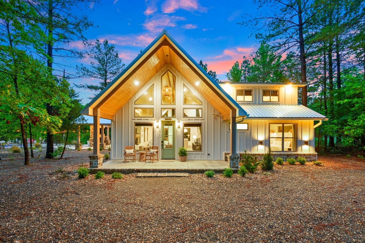 Welcome to our charming cottage - style retreat - glowing at twilight with a soft cream board and batten siding, a serene sage green door, cozy teak chairs for sipping morning coffee or tea.