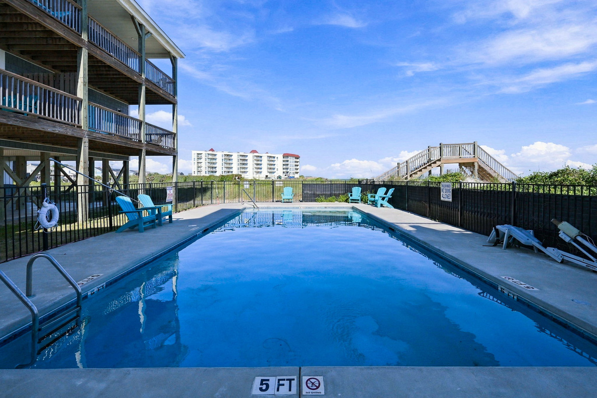 Ground-level view of the sparkling community pool enclosed by safety fencing, complete with poolside lounge chairs, step ladder entry, and a view of the private wooden dune crossover leading to the beach.   