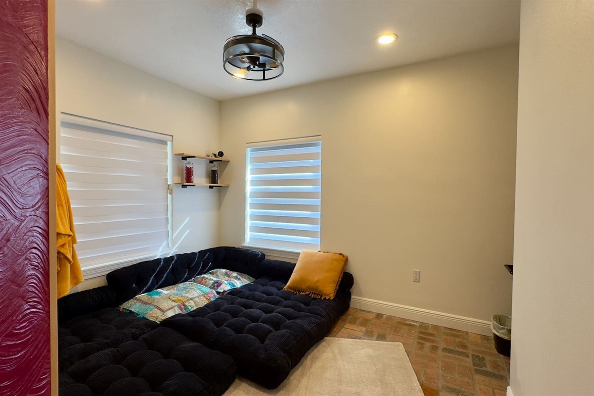 Cozy modern lounge with a low black sectional sofa, soft throw pillows, and natural light from dual windows with zebra blinds. Features tiled flooring, an area rug, and a ceiling fan with light for comfort.