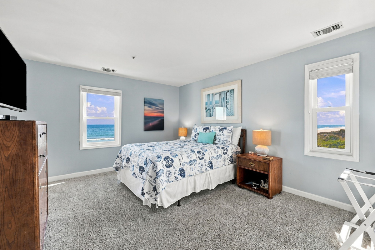 Bright guest bedroom featuring soft coastal blue walls, a bed dressed in ocean-themed patterned linens, warm wood furniture, and dual windows offering direct ocean views.   