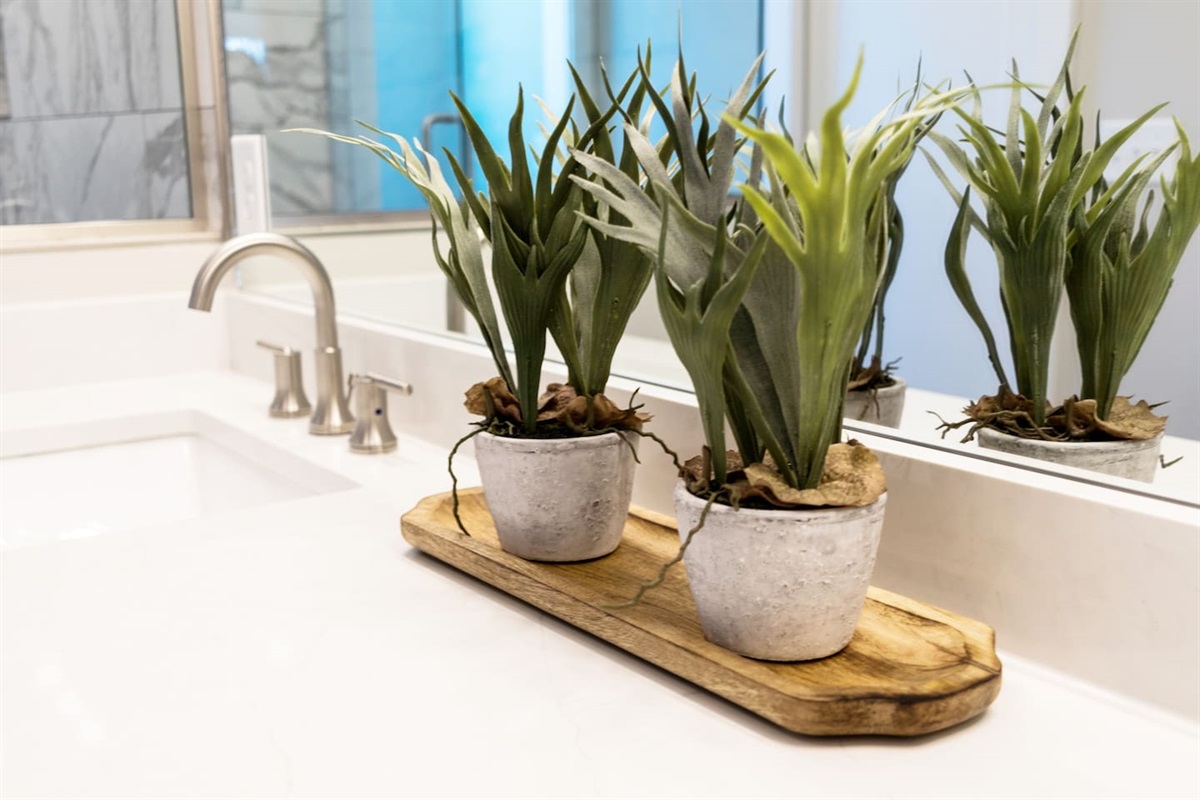 - Sleek countertop design with brushed nickel faucet and crisp white quartz surface - Twin faux succulents in rustic pots bring a fresh, natural touch to the bathroom décor - Wood tray base adds texture and warmth to the modern vanity arrangement