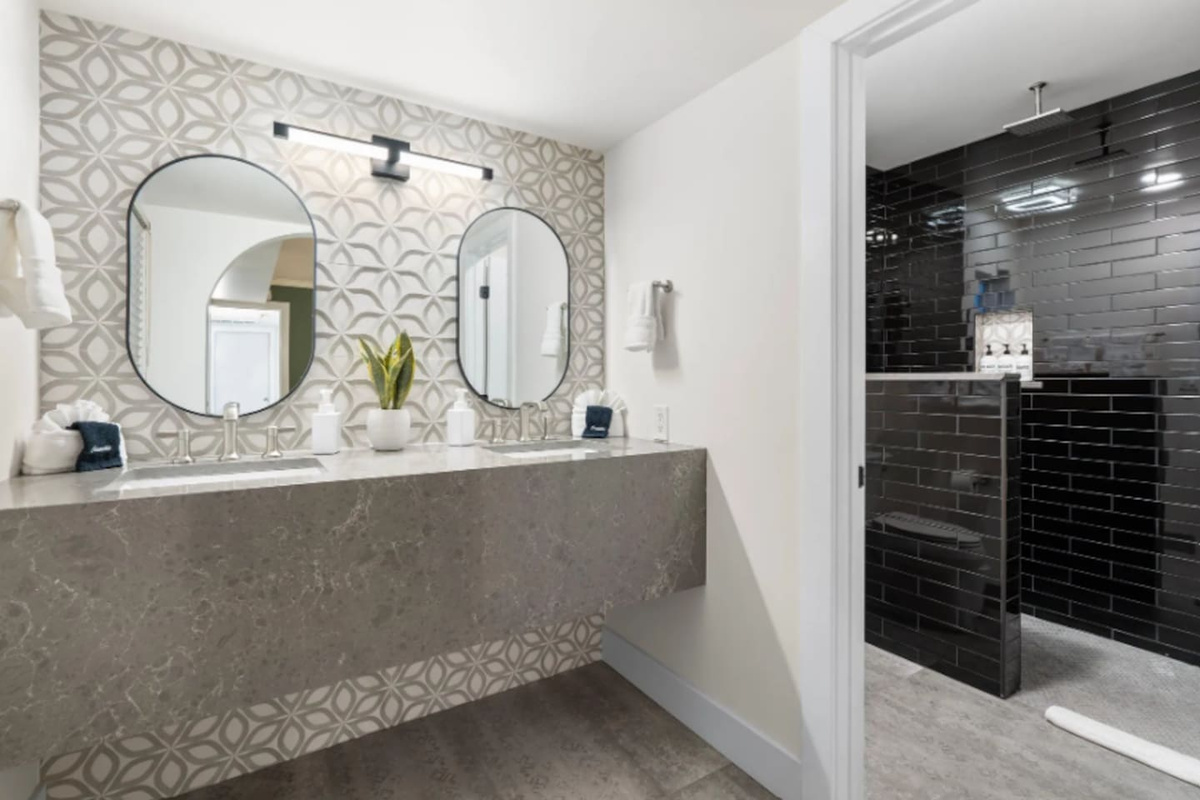 A dual vanity in patterned stone tile sets a spa-like tone, with a moody black-tiled shower just beyond. Every surface here feels intentional, right down to the oval mirrors and brushed fixtures.
