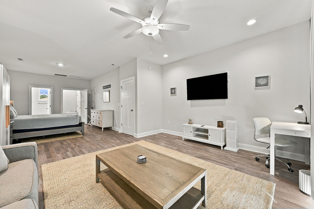 Bright, open bedroom suite view highlighting the combined layout with a king bed, living area with flat-screen TV, media console, work desk, and doorway access.   