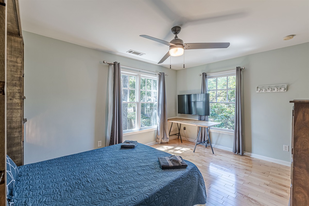 A sun-filled bedroom featuring a dresser, desk/table, TV, and blackout curtains for privacy and comfort.