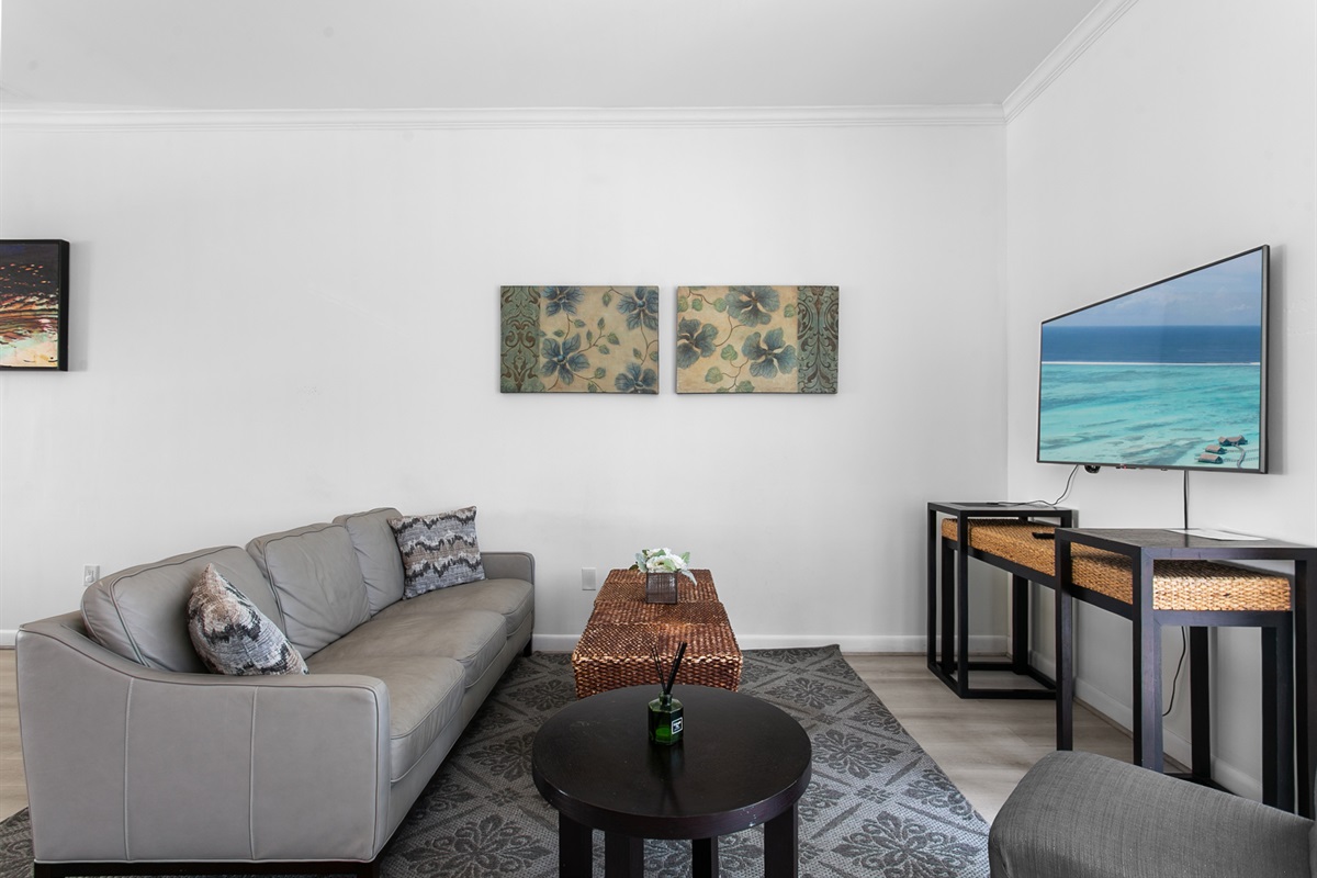 Living room with leather sofa, accent chairs, and a 50-inch flat screen TV. Sliding glass doors open to a Juliet balcony for sea breeze.