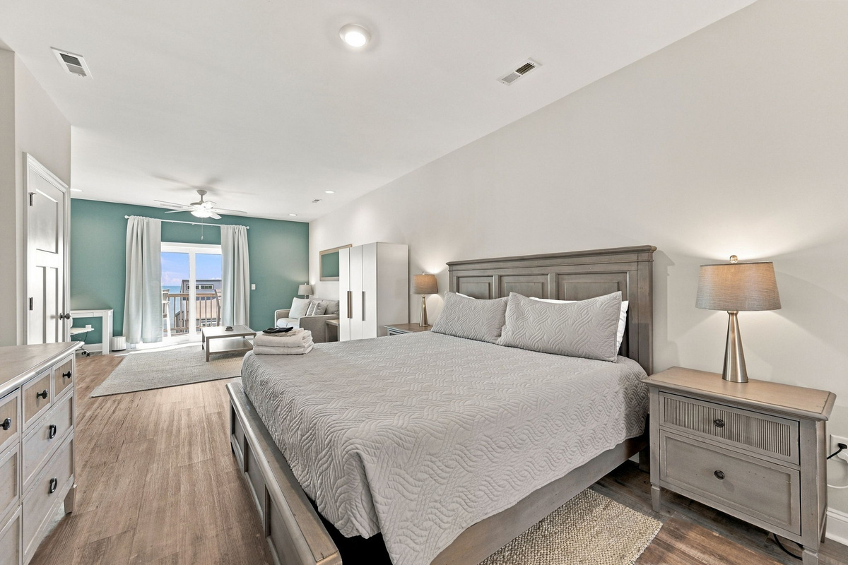 Spacious bedroom featuring a king bed with grey wooden furniture, neutral bedding, wood-look flooring, and an adjoining seating area with balcony access.   