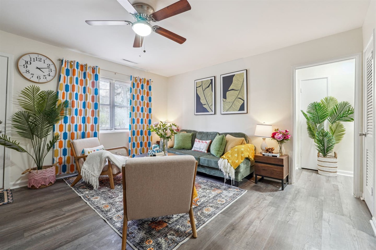 Enjoy a cozy living room featuring a long comfortable sofa, a TV for your entertainment, a ceiling fan, two chair, and a window with a lovely view outside — perfect for relaxing and unwinding.
