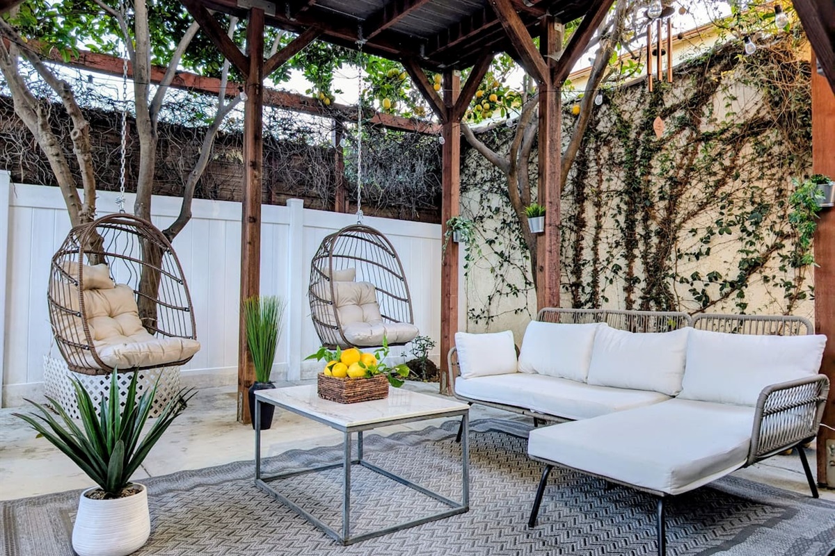This private, gated, and completely secluded covered patio offers total peace of mind with handy driveway access. A rustic, solid-roofed wooden pergola shelters a grill, sleek sectional, plush cushions, and twin hanging teardrop swings.